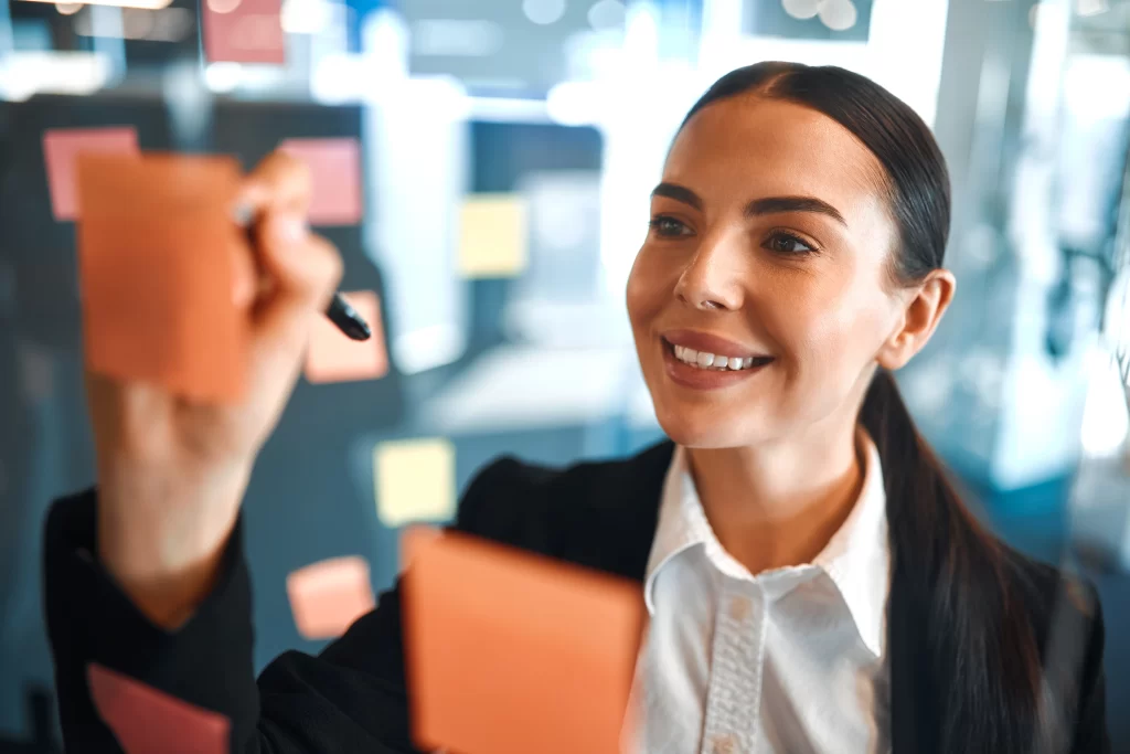 Colaboradora mulher morena dos olhos claros sorridente e profissional escrevendo no post it com uma caneta.