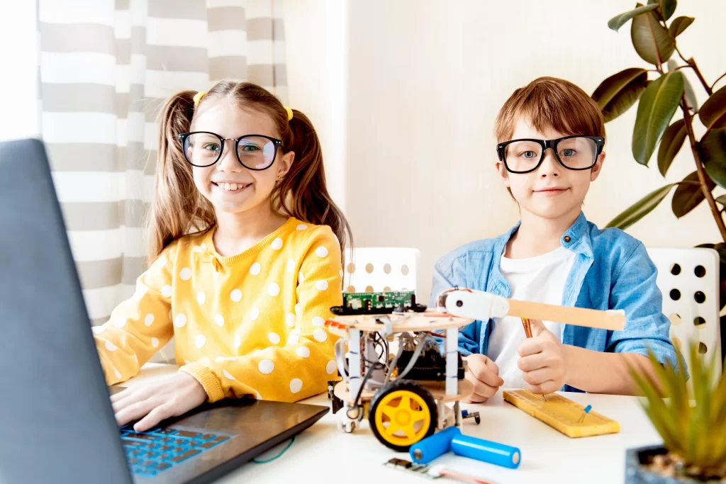 Crianças brancas utilizando óculos de grau, sorrindo ao estudarem junto da tecnologia através de notebook e também de um robô.