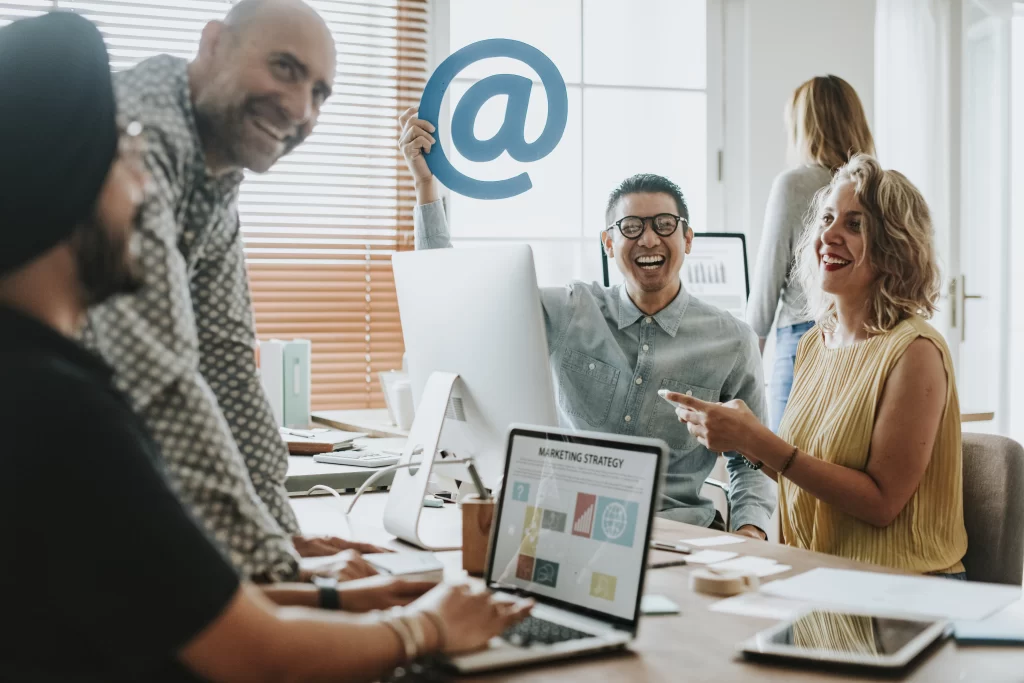 Cinco pessoas rindo e trabalhando juntos, sentados em uma mesa em volta de um notebook e um imac. Um dos integrantes sorridente segura em sua mão esquerda um arroba, é um homem branco que utiliza óculos de lentes de grau.
