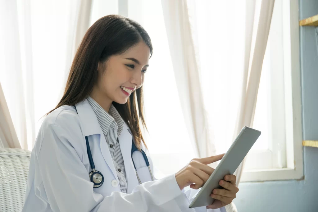 Mulher branca sorridente com um tablet na mão e um estetoscópio no pescoço em um ambiente hospitalar.