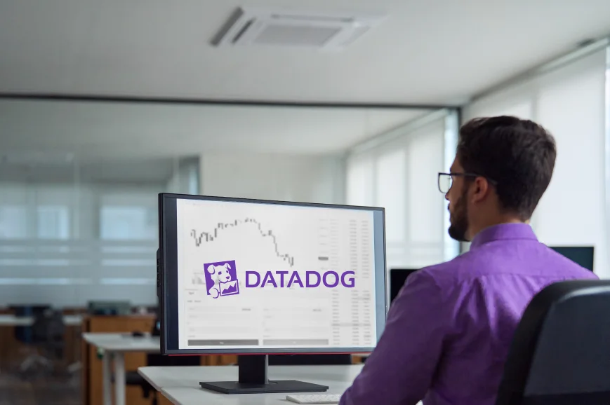 Homem branco de barba castanha e óculos, trabalhando, sentando em frente ao computador com a tela mostrando a logo da Datadog.